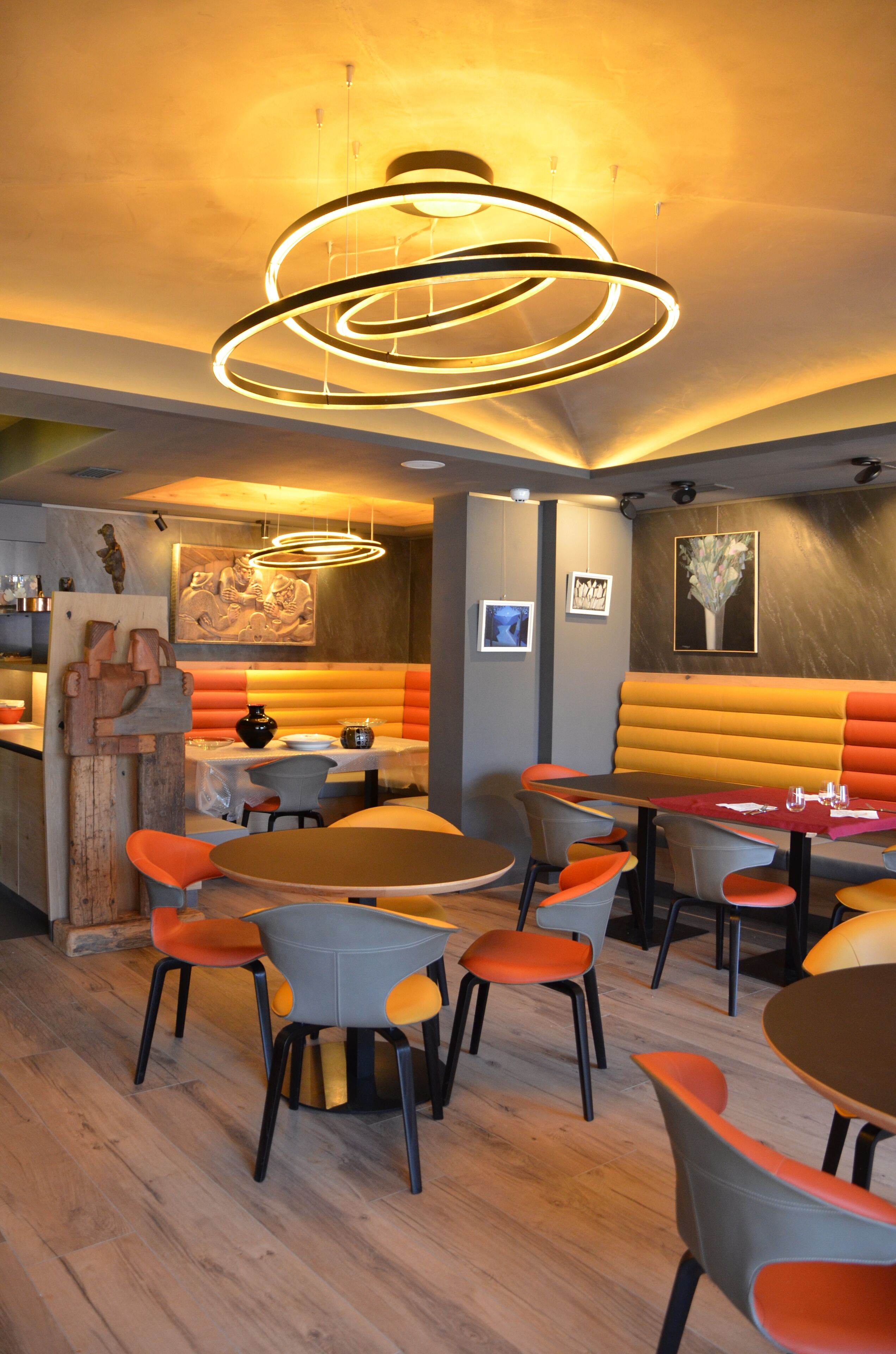 A restaurant interior with round tables, orange and grey chairs, a wooden bar area, and a modern pendant light fixture.