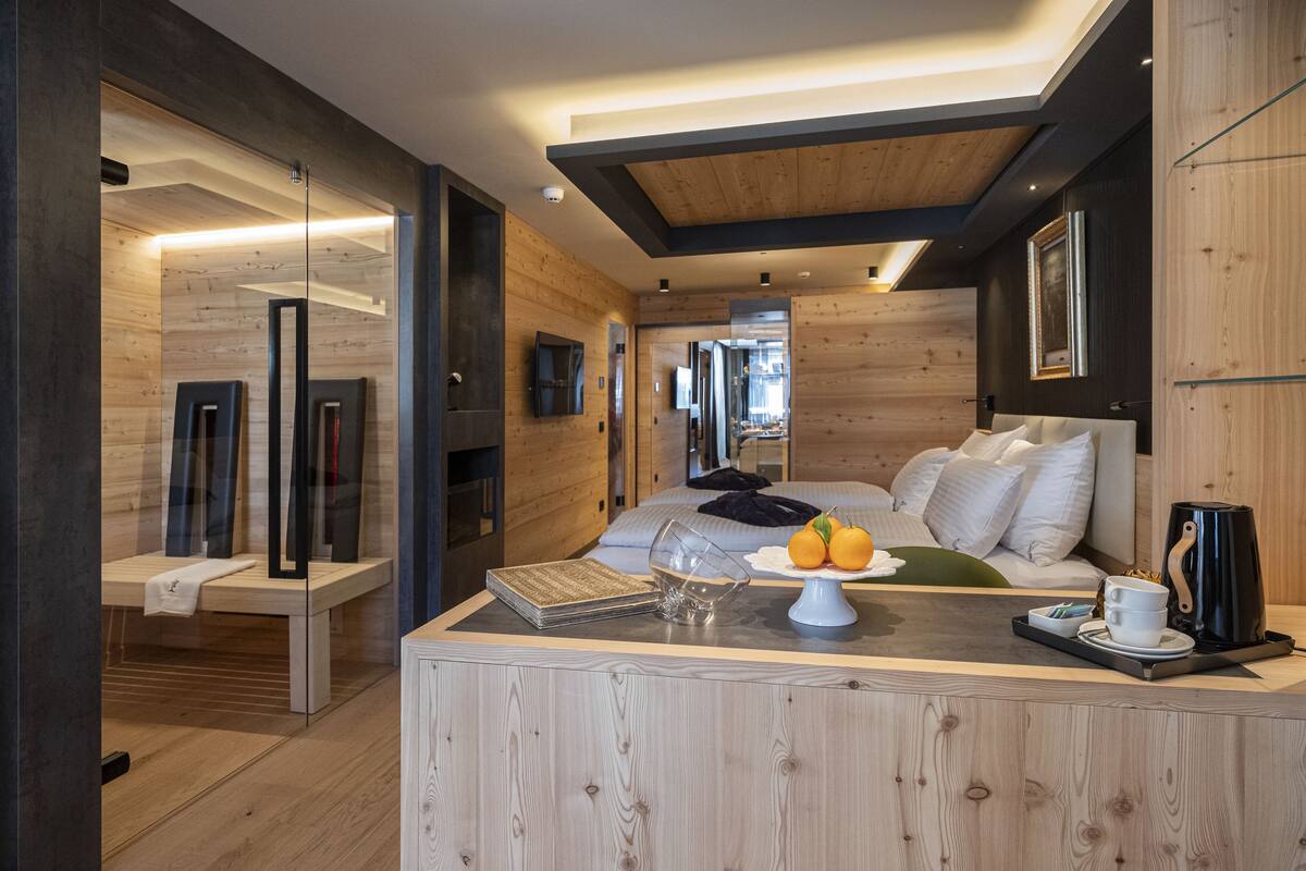 A modern bedroom with a wooden ceiling, a bed with white bedding, a bedside table with a coffee maker, and a view into the adjacent room.