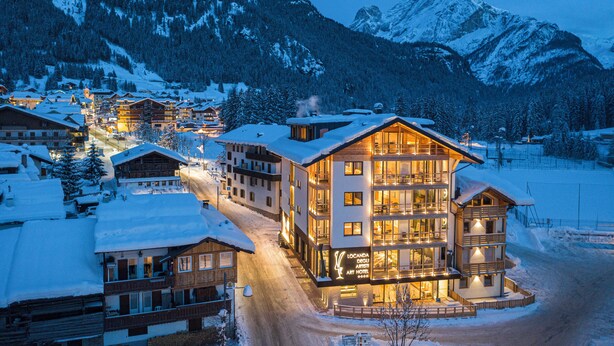 A snow-covered mountain town with a prominent hotel featuring multiple floors and balconies, surrounded by snow-covered trees and mountains. - Art Hotel Locanda degli Artisti (Canazei)