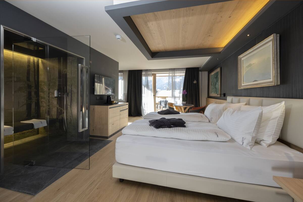 A modern hotel room with a large bed, a glass-enclosed shower, a wooden ceiling, and a view of the outdoors.