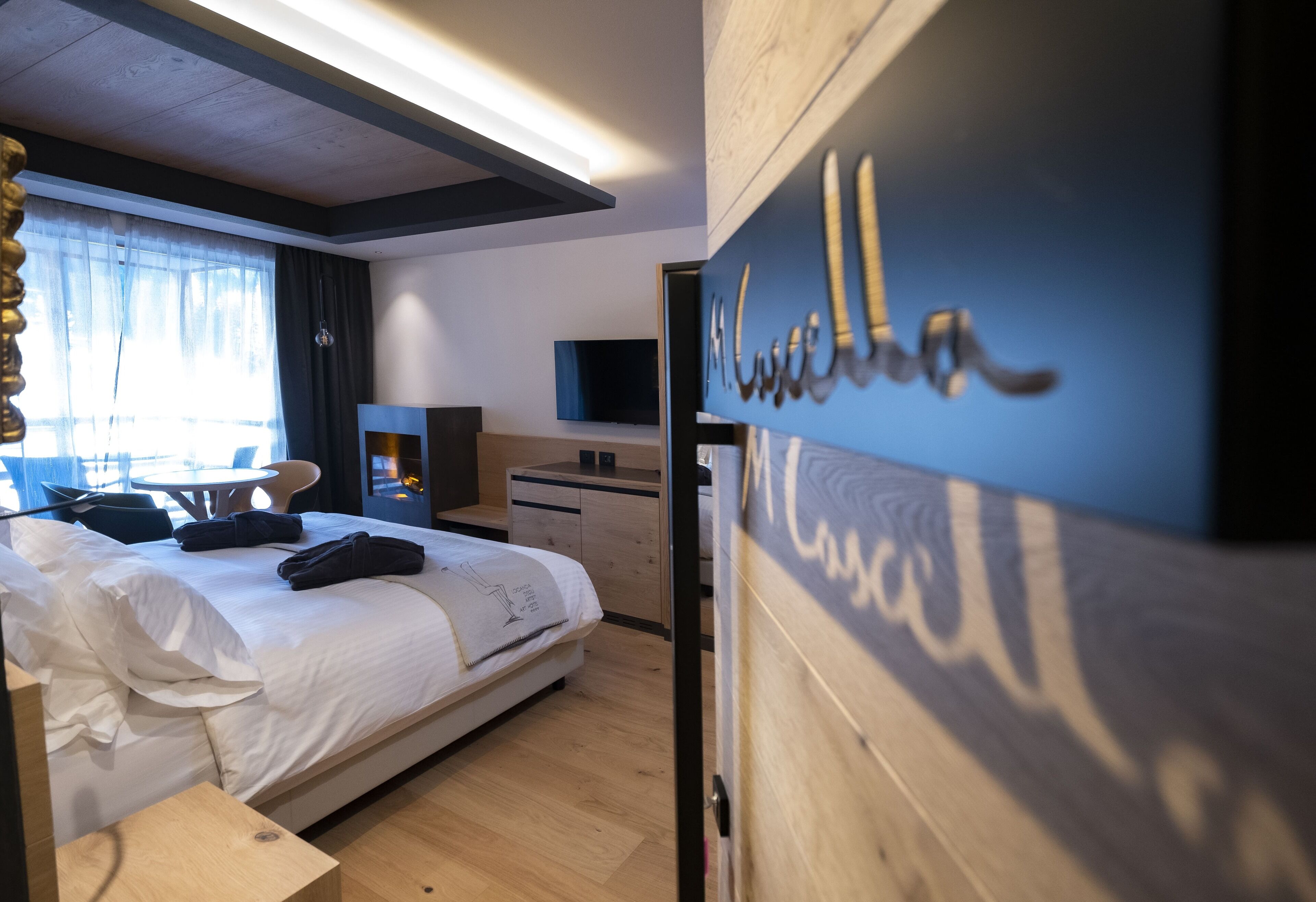 A hotel room with a bed, a desk, a chair, a TV, and a wooden wall with the name 'M. Louise'.