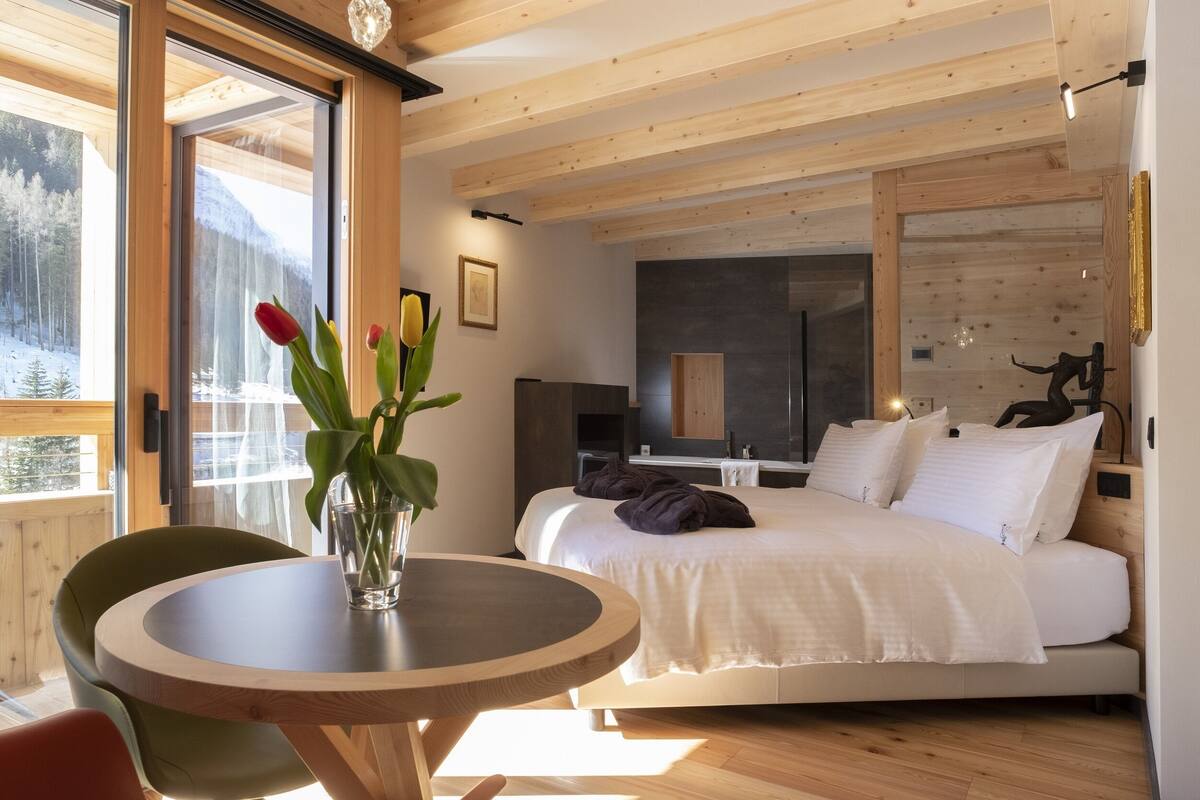 A modern bedroom with a large bed, a wooden ceiling, and a view of a snowy landscape through a large window.