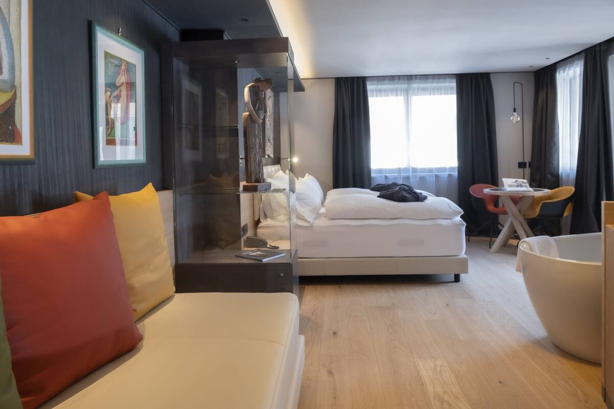 A modern hotel room with a bed, a seating area with a sofa and armchairs, a dining table, and artwork on the walls.