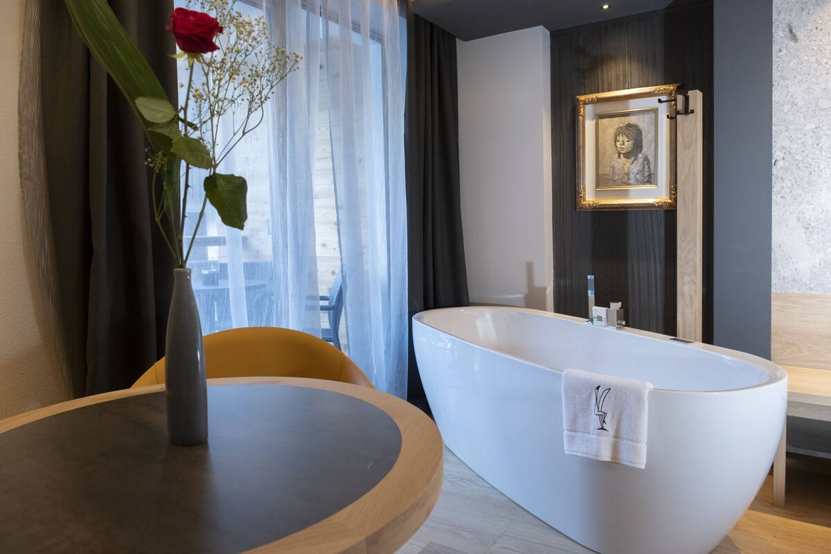 A modern bathroom with a freestanding bathtub, a round wooden table with a vase of flowers, and a framed picture on the wall.