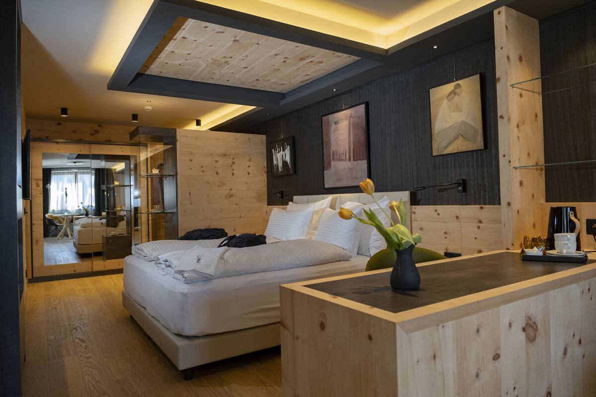 A modern hotel room with a wooden ceiling, a large bed, a bedside table, and a view into the adjacent living area.