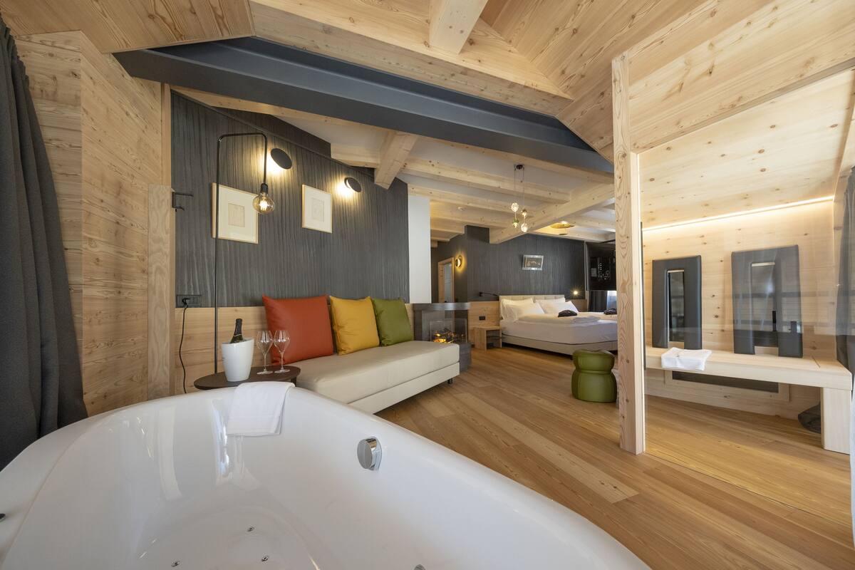 A modern hotel room with a large bathtub, a sofa with colorful cushions, a fireplace, and a separate bedroom area.