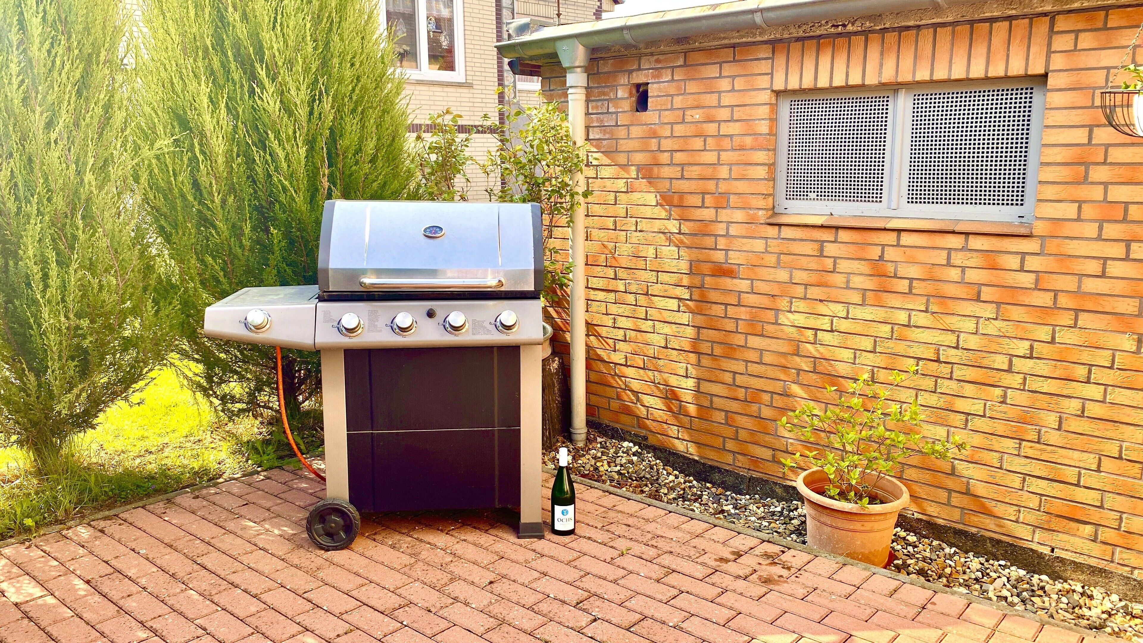A barbecue grill with four burners, a bottle of wine, and a potted plant in a garden area with a brick wall and a window.