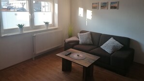 A living room with a dark brown sofa, a wooden coffee table with a remote control and a glass, and a window with blinds and a potted plant. - Double room at Ferienwohnung Kilian (Weberstedt)