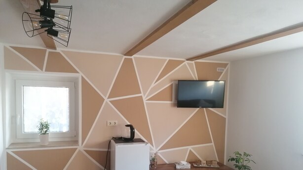 A modern living room with a geometric-patterned wall, a flat-screen TV mounted above a wooden console table, a small refrigerator, and a potted plant. - Double room at Ferienwohnung Kilian (Weberstedt)