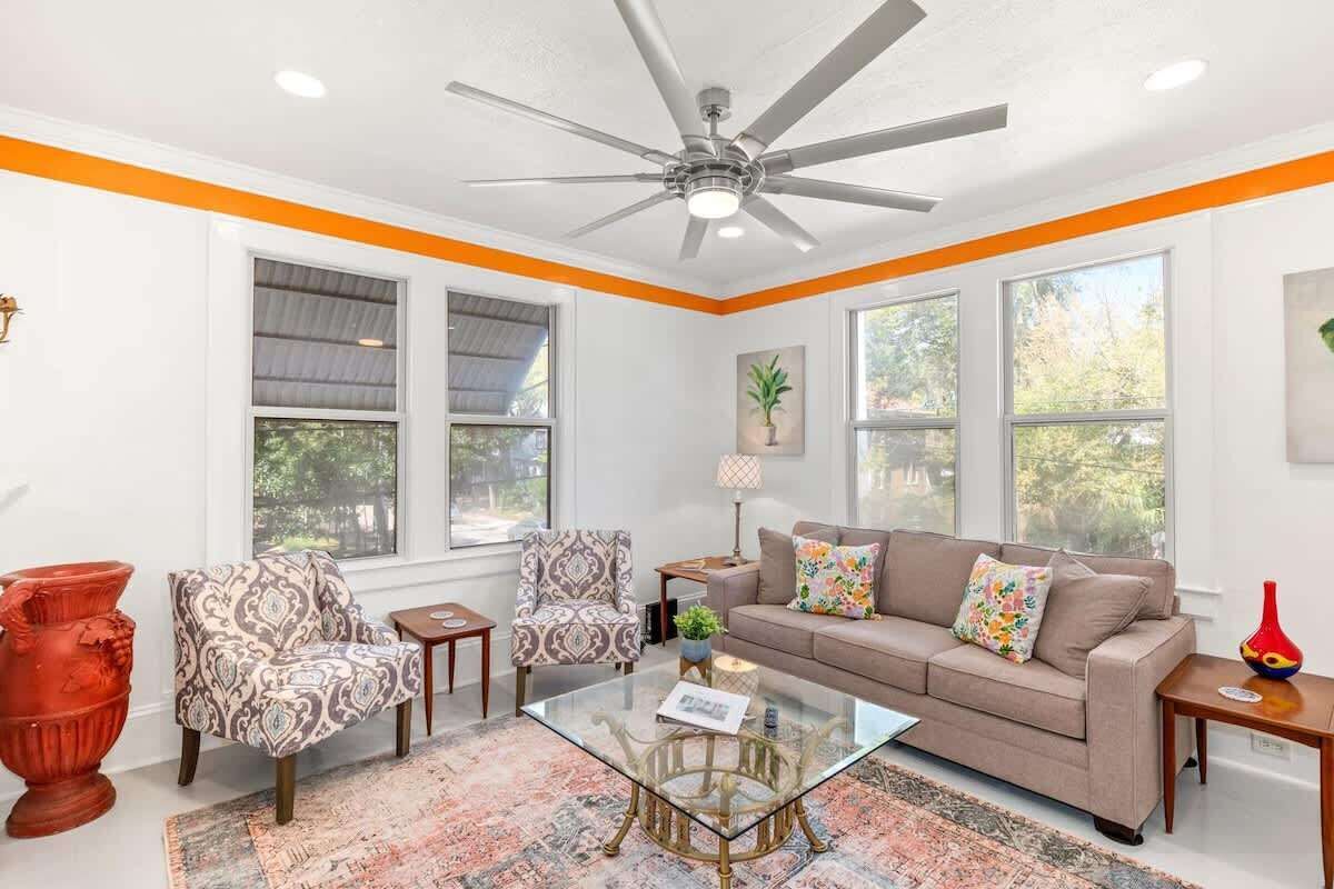 A living room with a beige sofa, patterned armchairs, a glass coffee table, and a ceiling fan.