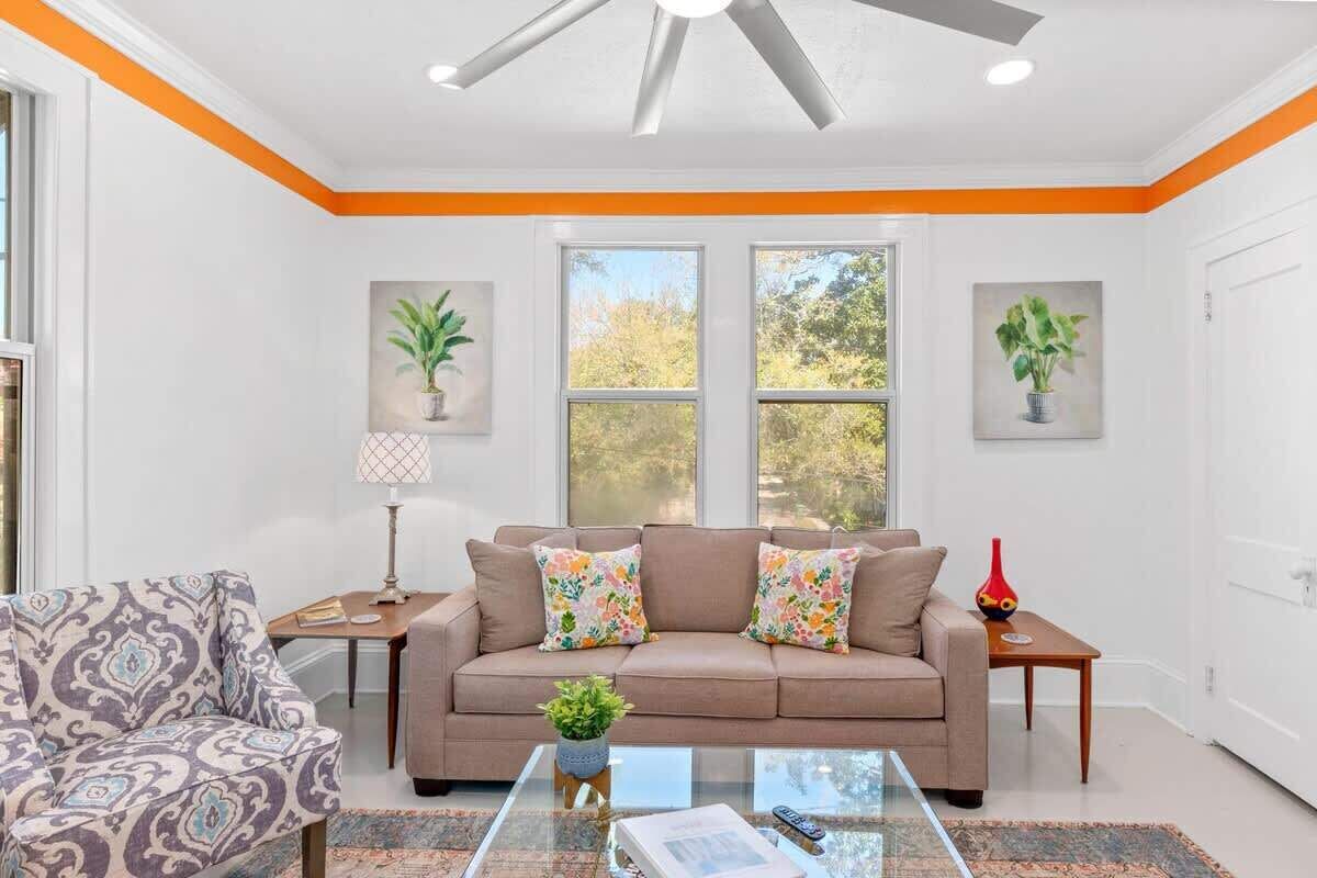 Una sala de estar con un sofá color beige, un sillón estampado y una mesa de centro de vidrio. Hay dos ventanas con adornos de plantas, un ventilador de techo y una puerta blanca.