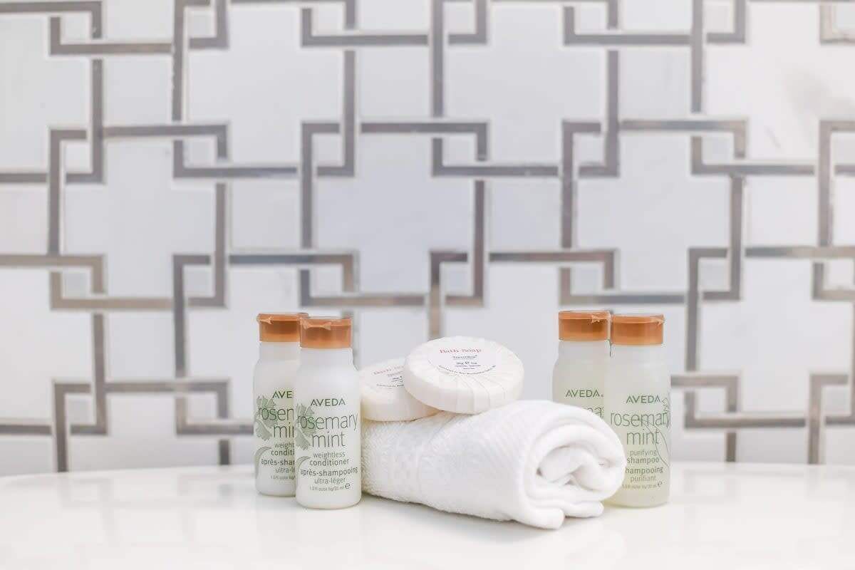 A bathroom with a geometric patterned wall, a white towel, and three bottles of Aveda rosemary mint shampoo.