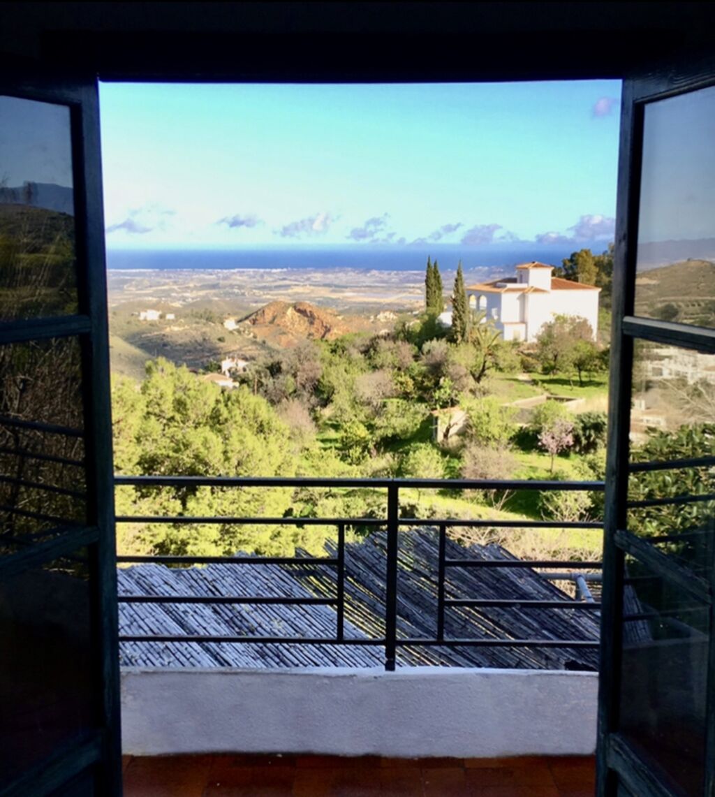 Une vue depuis un balcon doté d’une rambarde en métal, donnant sur un paysage verdoyant, quelques bâtiments et des montagnes lointaines, le tout sous un ciel dégagé.