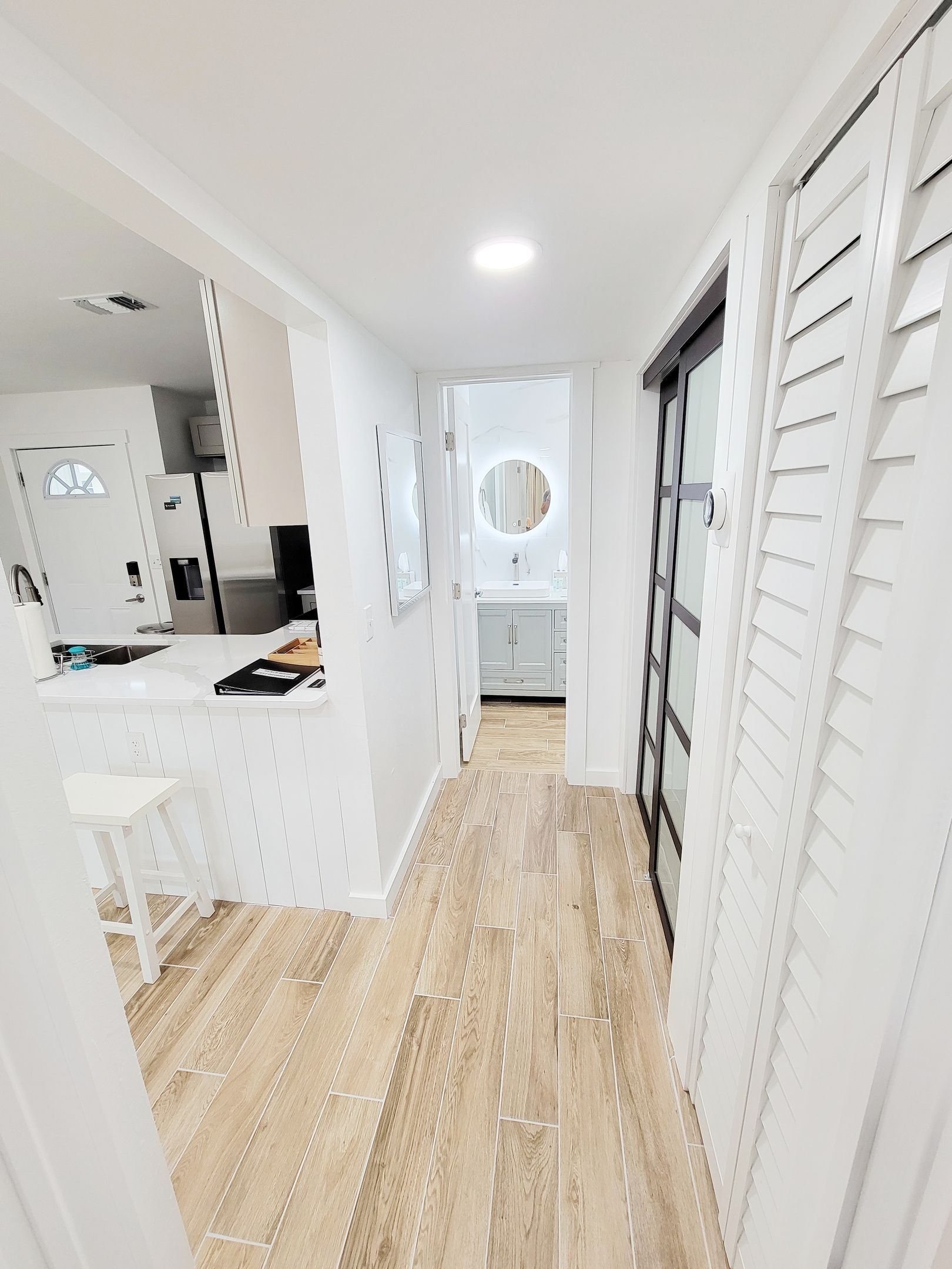 A modern kitchen with white cabinetry and a central island, a hallway with wooden flooring, and a view into a bathroom with a round mirror.