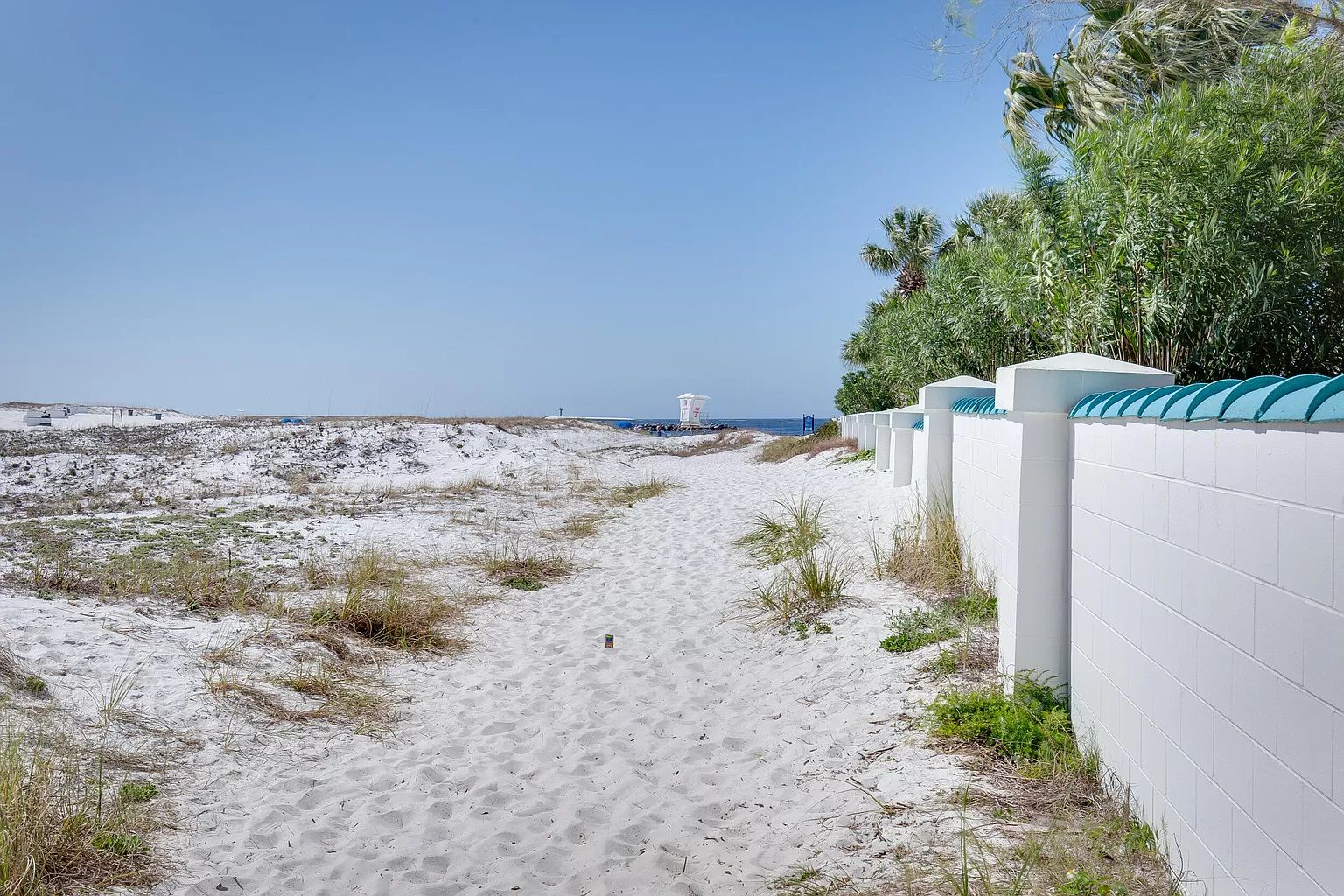 Een zandpad langs het strand dat naar de oceaan leidt, met een witte muur en begroeiing aan de zijkant.