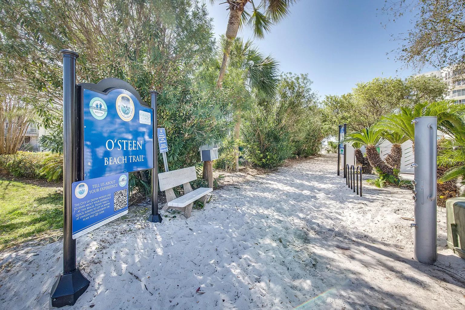 Señal de sendero en la playa con código QR para comentarios, un banco y un camino arenoso con árboles.