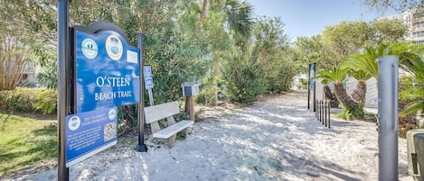 Un panneau indiquant un sentier menant à la plage, avec un code QR pour recueillir les commentaires des visiteurs, un banc et un chemin de sable bordé d’arbres.