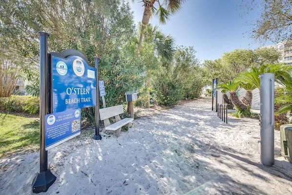 A beach trail sign with QR code for feedback, a bench, and a sandy path with trees.