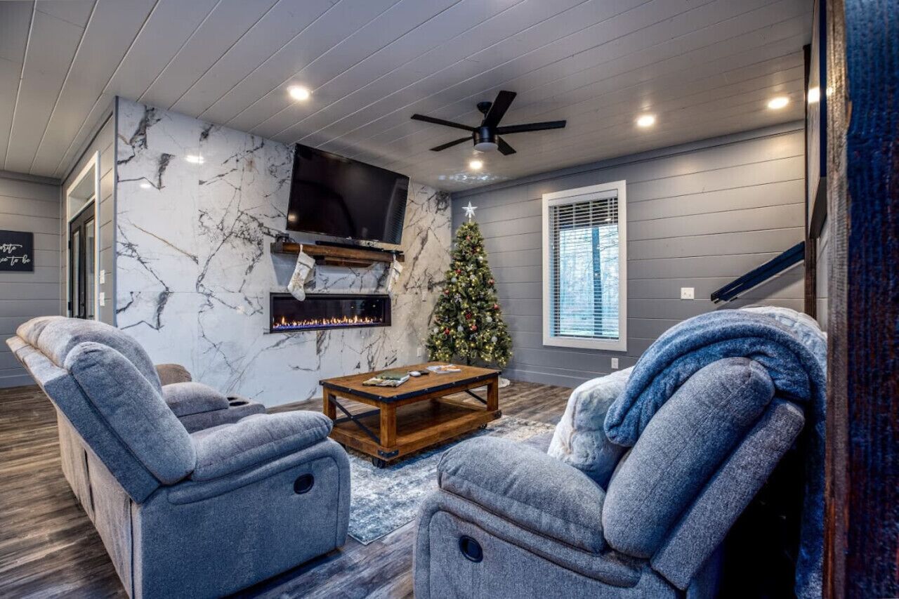 A living room with a fireplace, a TV mounted on the wall, a Christmas tree, and grey upholstered chairs.