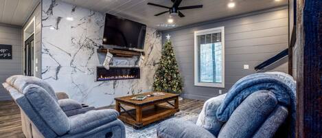 A living room with a fireplace, a TV mounted on the wall, a Christmas tree, and grey upholstered chairs.