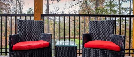 A balcony with two wicker chairs and a glass table, overlooking a forested area.
