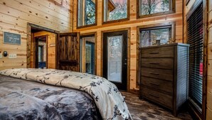 A log cabin bedroom with a bed, dresser, and a window with a view of trees. - Dawn's Early Peak - Modern cabin w/ birds-eye view (Smithville)