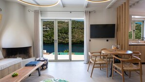Een moderne woonkamer met een open haard, een eethoek met houten stoelen, een flatscreen televisie en uitzicht op een meer en heuvels.