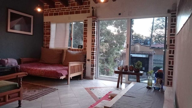 Una habitación acogedora con una cama, una silla de madera, una mesa con un florero y un ventanal corredero que da a un espacio exterior.