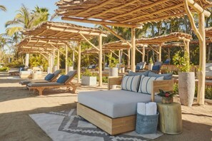 A beachside lounge area with wooden loungers and a sofa under thatched umbrellas, surrounded by palm trees.