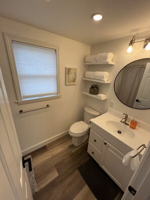 Une salle de bain moderne avec des toilettes, un lavabo et un miroir. On y trouve des serviettes sur des étagères et une fenêtre avec des stores.