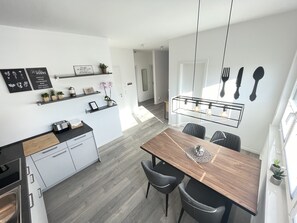 A modern kitchen with white cabinets, a wooden countertop, and a dining area with a wooden table and chairs. There are shelves with plants and kitchen utensils, and a hanging light fixture above the table.