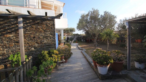 Une terrasse en bois menant à un mur de pierre, entourée de plantes en pot et d’un bâtiment doté d’un balcon.