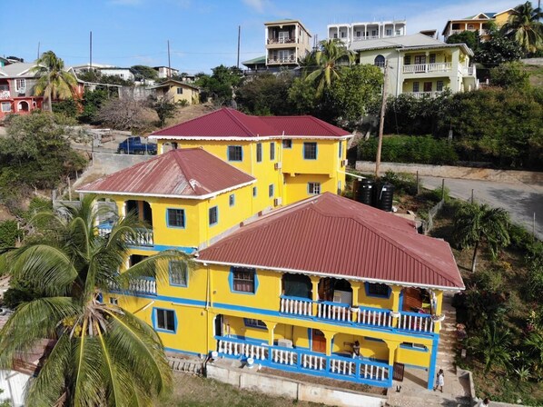 Un edificio amarillo de dos pisos con techo rojo, un balcón y un paisaje circundante con palmeras y otros edificios.