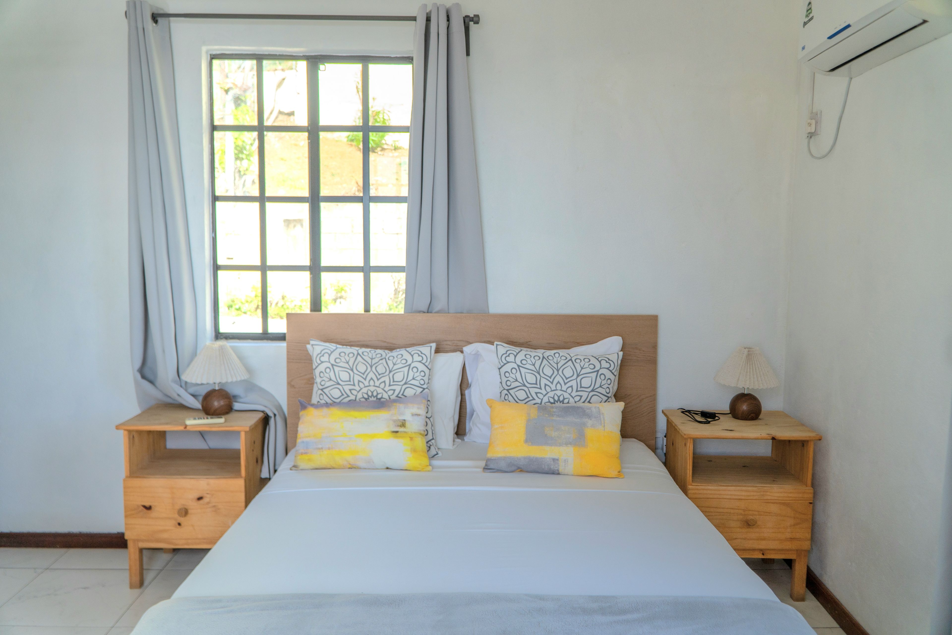 Une chambre avec un grand lit, deux tables de chevet, une fenêtre avec des rideaux et un appareil de climatisation.