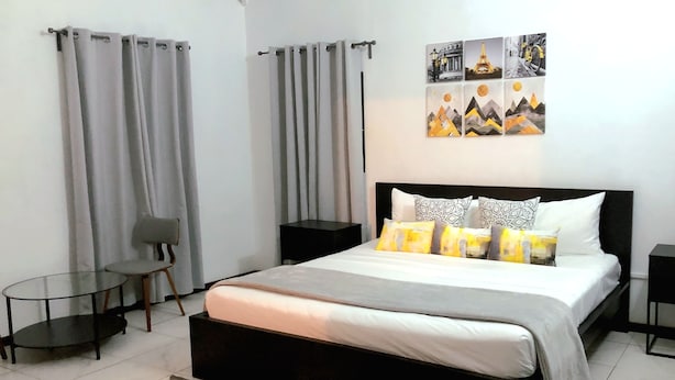 Een moderne slaapkamer met een zwart bed, een grijs-wit dekbed en gestreepte kussens. Er staat een klein rond tafeltje met een stoel, een zwart nachtkastje en een wandplank.