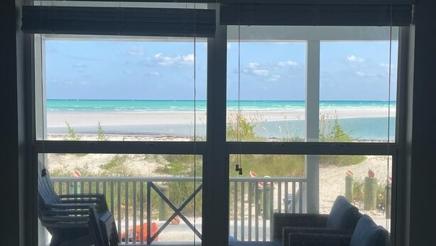 A room with a large window overlooking a beach. There are two chairs and a small table on a wooden deck. - Sandy & Salty Beach Cottage (Spanish Wells)