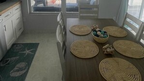 A dining area with a view of the beach, a wooden table with placemats, chairs, and a small bowl of decorative items. - Sandy & Salty Beach Cottage (Spanish Wells)