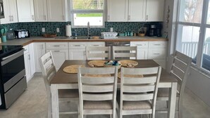 A kitchen with white cabinets, a wooden dining table, and a window with a view of greenery. - Sandy & Salty Beach Cottage (Spanish Wells)