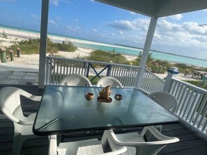 Une table à plateau en verre, entourée de chaises en plastique blanc, sur une terrasse en bois donnant sur une plage.
