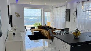 A modern kitchen with a central island, a dining area with a table and chairs, and a sofa facing a large window with a view of the sea. - VILLA PHARE CARAIBES (Le Moule)