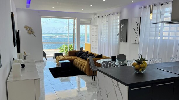 A modern kitchen with a large island, a dining area with a sofa, and a view of the ocean. - VILLA PHARE CARAIBES (Le Moule)