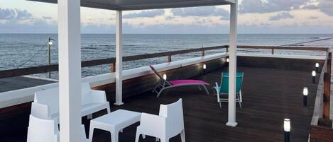 A covered outdoor area with a wooden deck, white furniture, and a view of the ocean.