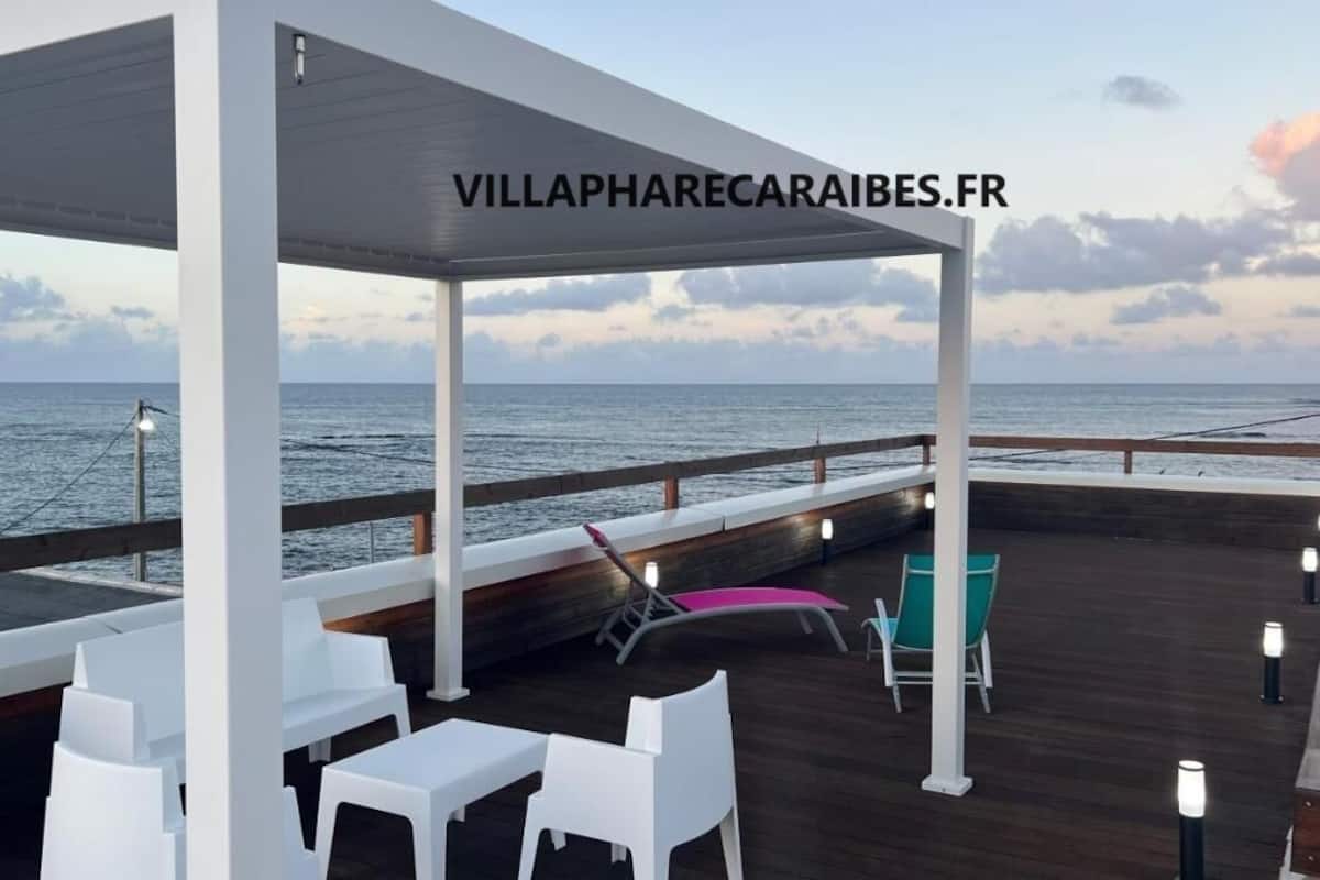 A covered outdoor area with wooden flooring, white tables, and chairs, overlooking the sea.