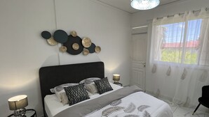 A bedroom with a bed, a black chair, a window with sheer curtains, and wall art. - VILLA PHARE CARAIBES (Le Moule)