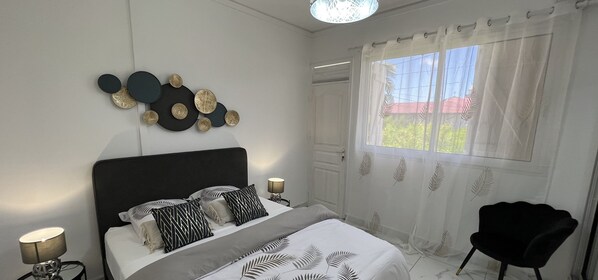 A bedroom with a bed, a black chair, a window with sheer curtains, and wall art. - VILLA PHARE CARAIBES (Le Moule)