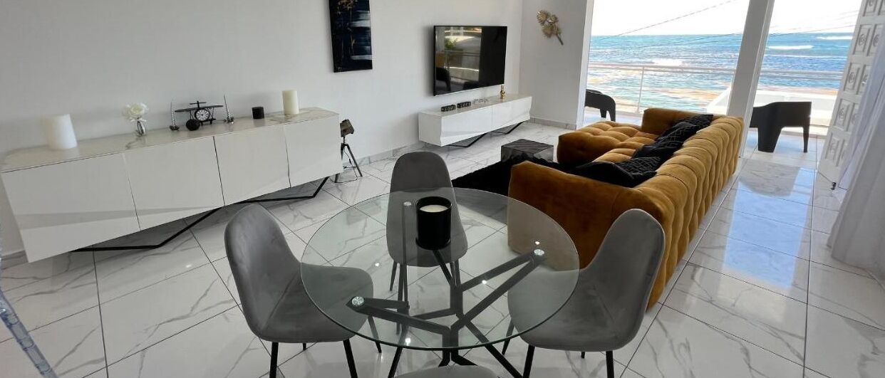 A modern living room with a glass dining table, grey chairs, a mustard-colored sofa, a white entertainment unit, and a large TV.