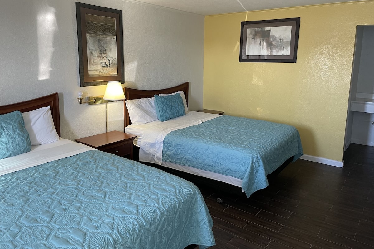A hotel room with two beds, a nightstand with a lamp, a framed picture on the wall, and a wooden floor.