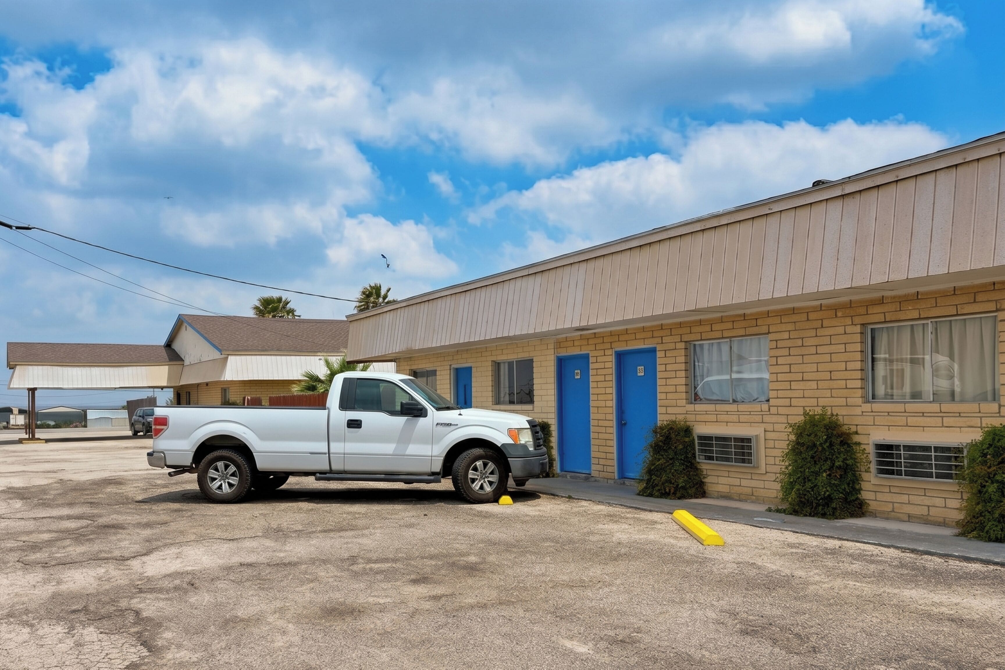 A motel with a white pickup truck parked in front, a row of blue doors, and a clear sky.