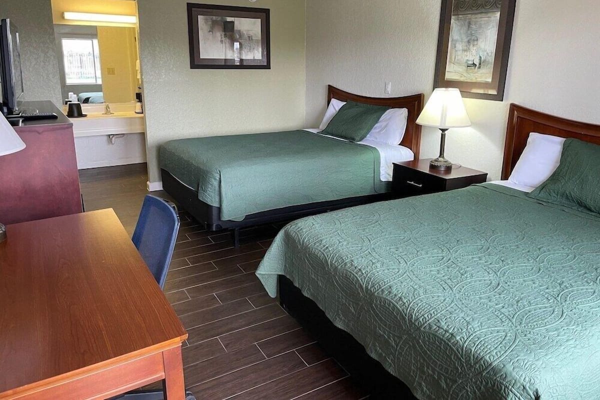 A hotel room with two beds, a desk, a chair, a ceiling fan, and a bathroom visible through an open door.
