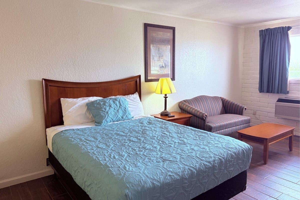 A hotel room with a bed, a chair, a table, a lamp, and a picture on the wall.
